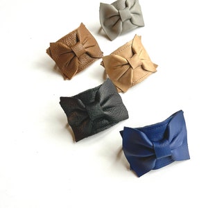 May include: Five leather bow hair clips in different colors: gray, brown, beige, black, and blue.