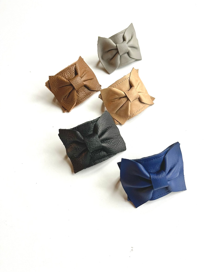 May include: Five leather bow hair clips in different colors: gray, brown, beige, black, and blue.