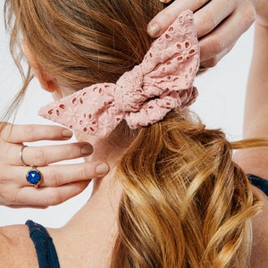 May include: A pink crocheted hair scrunchie with a bow. The scrunchie is tied around a woman's ponytail.