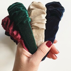 May include: Four velvet headbands in different colors: burgundy, green, beige, and navy blue. The headbands are gathered and bunched together in a hand.
