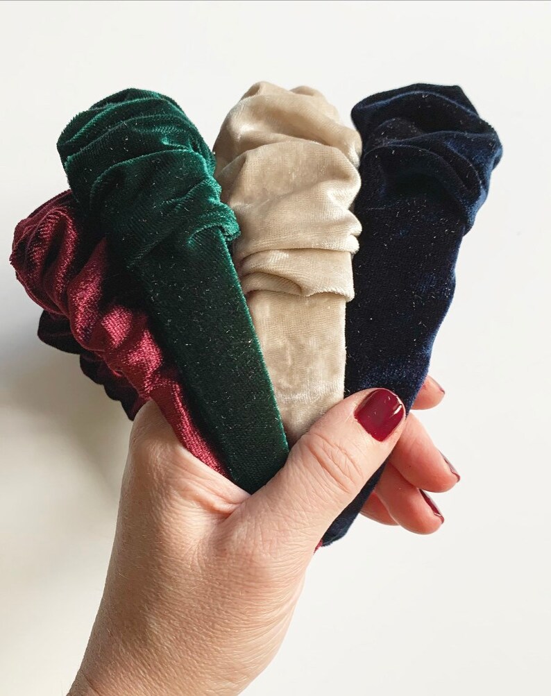 May include: Four velvet headbands in different colors: burgundy, green, beige, and navy blue. The headbands are gathered and bunched together in a hand.