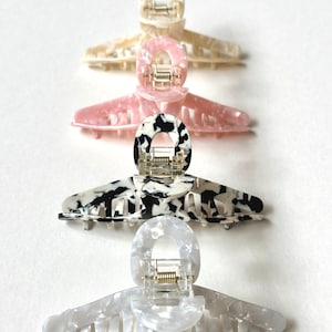 Puede incluir: Cuatro pinzas para el pelo de diferentes colores y patrones. Las pinzas están hechas de un material plástico translúcido y tienen una forma curva con un mecanismo de resorte. Los colores son blanco, rosa, blanco y negro, y blanco con un efecto marmoleado.