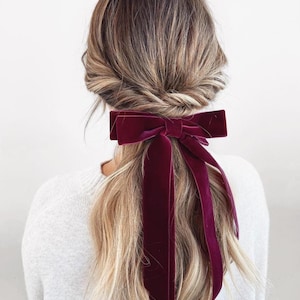 May include: A burgundy velvet hair bow tied in a ponytail. The bow is large and rectangular, with long, flowing ribbons. The hair is blonde and styled in a loose updo. The person is wearing a white sweater.