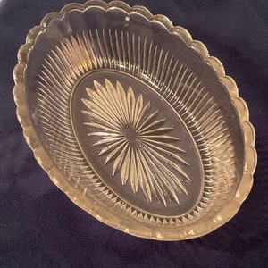 May include: Clear glass oval serving dish with a scalloped edge. The dish features a starburst design in the center and vertical lines radiating from the center. The dish is made of clear glass.