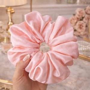 May include: A large, pale pink satin scrunchie. The scrunchie is made of soft, gathered fabric and designed for hair. The colour is a delicate pink, and the fabric has a subtle sheen. A hair accessory.