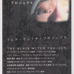 May include: A promotional card for the film "The Blair Witch Project", featuring Japanese and English text. The card has a dark, textured background and includes the film's title, credits, and a partial facial image.