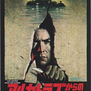 Könnte beinhalten: Ein Filmplakat für den Film "Flucht aus Alcatraz" mit Clint Eastwood. Das Plakat zeigt eine Nahaufnahme von Eastwoods Gesicht, das durch eine rissige Wand lugt und ein Messer hält. Der Text auf dem Plakat lautet "Escape from Alcatraz" und "Clint Eastwood".