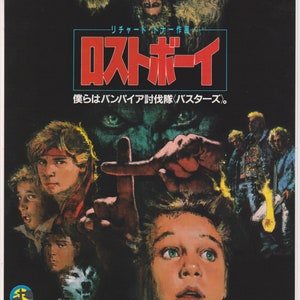 May include: A movie poster for the film "The Lost Boys" featuring a group of young men and a vampire. The poster is in Japanese and features the text "Lost Boys" in red letters and the text "We are the vampire hunters" in smaller letters below. The poster also features a cross and a vampire with glowing red eyes.