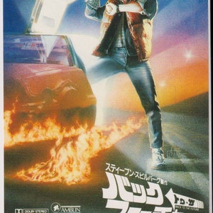May include: A movie poster for the film Back to the Future, featuring a young man in a vest and jeans standing in front of a DeLorean time machine. The poster is red and white with the title "Back to the Future" in large, bold letters. The poster also includes the names of the actors, director, and producers.