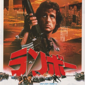 May include: A movie poster for the film "First Blood" starring Sylvester Stallone. The poster features a red and white title card with the Japanese word for "Rambo" in large, bold letters. The poster also features a photo of Sylvester Stallone holding a rifle, with a cityscape in the background. The poster text reads: "'83 New Year (Action Big) excitement finally reaches a huge climax!" and "Now, the fierce battle begins!" The poster also includes the Japanese text: "December 18th, "White Heat" Roadshow"
