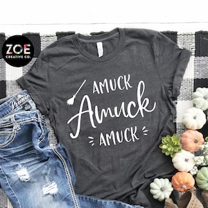 May include: A dark grey t-shirt with the text "AMUCK Amuck AMUCK=" in white. The shirt is laying on a black and white checkered rug with a pair of brown cut-out ankle boots and small decorative pumpkins.