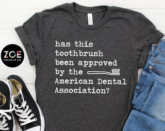 May include: A dark grey t-shirt with the text "has this toothbrush been approved by the American Dental Association?" and a graphic of a toothbrush. The t-shirt is displayed on a white wooden surface with a pair of black sneakers and blue jeans.