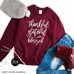 May include: Maroon sweatshirt with the text "thankful grateful blessed" in white lettering. The sweatshirt is lying on a pair of blue jeans and a maroon hat.