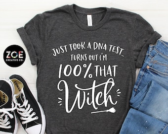 May include: A dark gray t-shirt with the text "Just took a DNA test... Turns out I'm 100% that Witch" in white lettering. The shirt is paired with blue jeans and black sneakers.