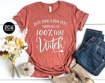 May include: A rust-colored t-shirt with the text "Just took a DNA test. Turns out I'm 100% that Witch" in white lettering. The shirt is laid out on a white wooden surface with a bouquet of flowers, a small pumpkin, and a pair of jeans.