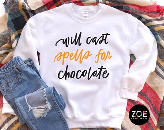 May include: White sweatshirt with the text "Will cast spells for chocolate" in black and orange lettering. The sweatshirt is laid out on a plaid blanket with a pair of blue jeans.