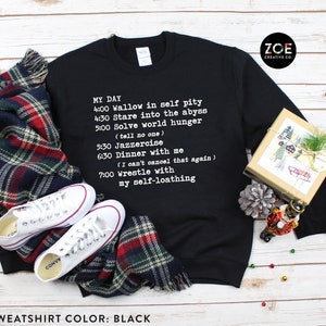 May include: Black unisex sweatshirt with a humorous daily schedule printed in white. The image shows the sweatshirt alongside white Converse trainers, a tartan scarf, and a gift box. The Zoe Creative Co. logo is visible.