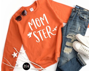 May include: Orange sweatshirt with the text "MOM *STER" in white with a ghost and bat graphic. The sweatshirt is laid flat on a white surface with a pair of sunglasses, a pair of jeans, and white sneakers.