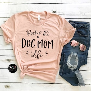 May include: A peach colored t-shirt with the text "Rockin' the DOG MOM Life" in black lettering. The shirt is folded and laid on a white wooden surface with a pair of blue jeans and sunglasses.