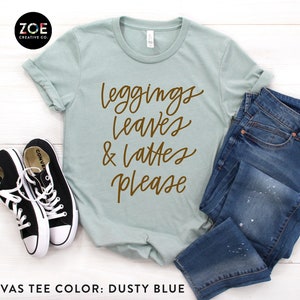 May include: A dusty blue Bella Canvas t-shirt with the text "leggings leaves & lattes please" in brown lettering.