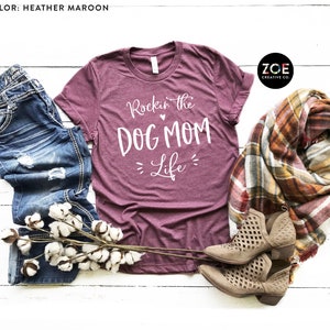 May include: Heather maroon t-shirt with white text that reads "Rockin' the Dog Mom Life". The shirt is displayed on a white wood background with a pair of brown ankle boots, a plaid scarf, and a sprig of cotton.
