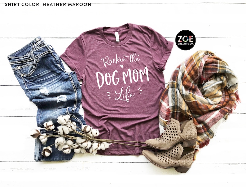 May include: Heather maroon t-shirt with white text that reads "Rockin' the Dog Mom Life". The shirt is displayed on a white wood background with a pair of brown ankle boots, a plaid scarf, and a sprig of cotton.