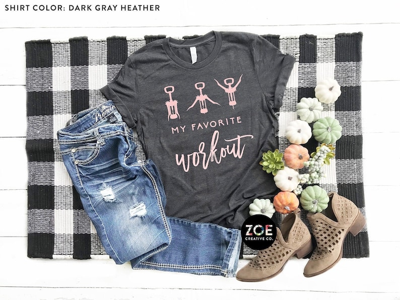 May include: A dark gray heather t-shirt with a graphic of three wine openers in different poses. The text "My Favorite Workout" is written in pink below the graphic. The shirt is laid out on a black and white checkered rug with a pair of brown cut-out ankle boots and blue jeans.