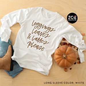 May include: White long-sleeve shirt with brown text that reads "leggings, leaves & lattes please". The shirt is laid flat on a wood surface with a pumpkin and fall leaves.