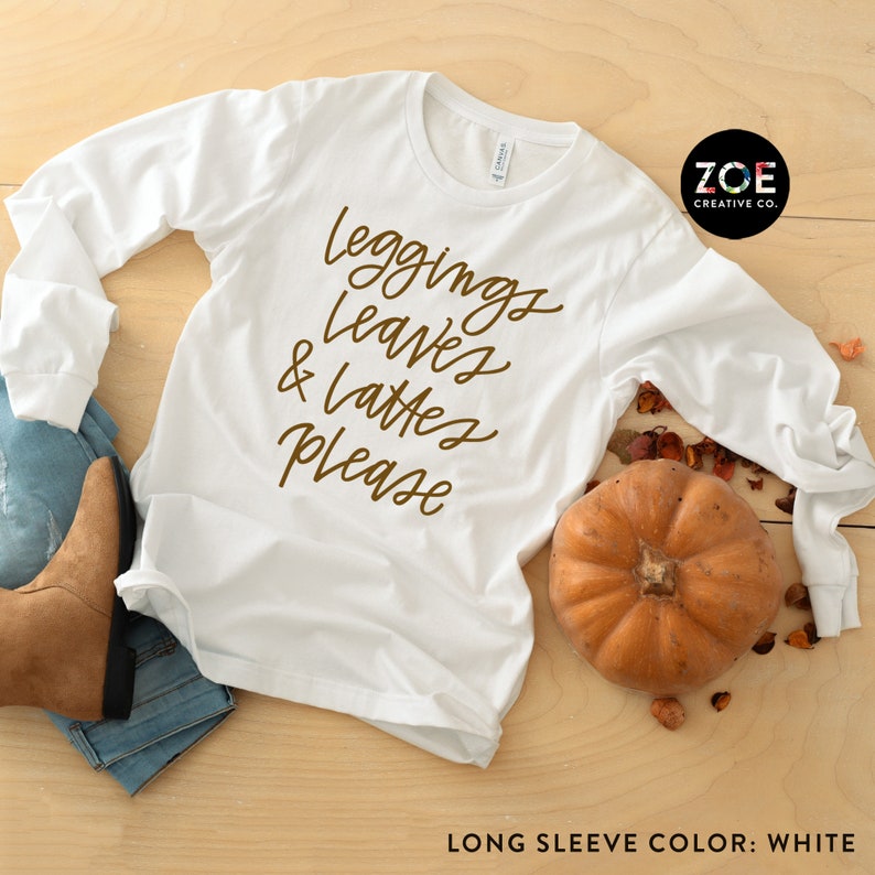 May include: White long-sleeve shirt with brown text that reads "leggings, leaves & lattes please". The shirt is laid flat on a wood surface with a pumpkin and fall leaves.