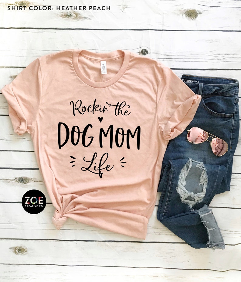 May include: A peach colored t-shirt with the text "Rockin' the DOG MOM Life" in black lettering. The shirt is folded and laid on a white wooden surface with a pair of blue jeans and sunglasses.