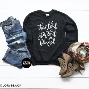 May include: Black sweatshirt with the text "thankful grateful blessed" in white lettering. The sweatshirt is laid flat on a white surface with a pair of blue jeans, a scarf, and brown boots.