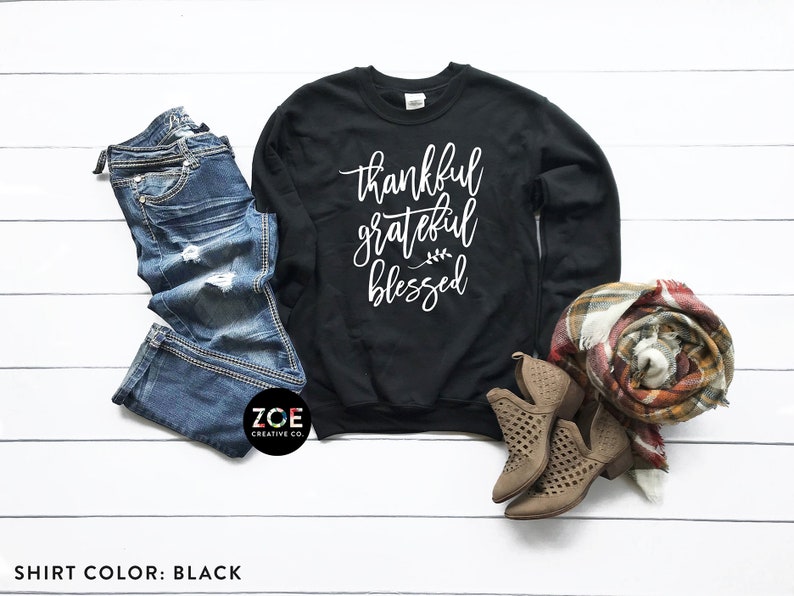 May include: Black sweatshirt with the text "thankful grateful blessed" in white lettering. The sweatshirt is laid flat on a white surface with a pair of blue jeans, a scarf, and brown boots.