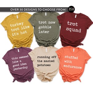 May include: Six different t-shirts with Thanksgiving-themed text printed in white on various coloured shirts. The shirts are folded and arranged in a grid pattern. The text on the shirts includes "turkey trot like it's hot", "trot now gobble later", "trot squad", "this seemed like a good idea yesterday", "running off the mashed potatoes", and "stuffed with endurance".