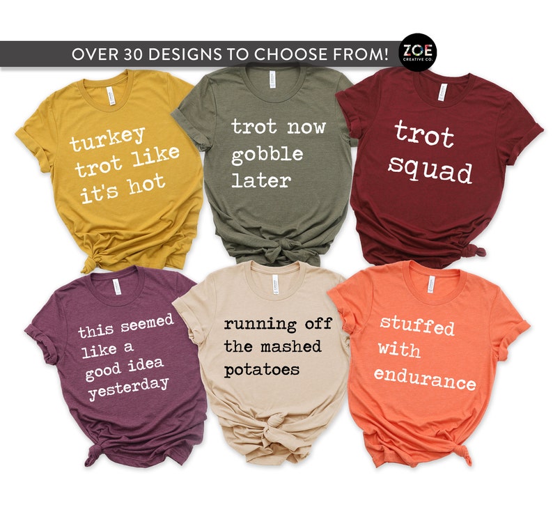 May include: Six different t-shirts with Thanksgiving-themed text printed in white on various coloured shirts. The shirts are folded and arranged in a grid pattern. The text on the shirts includes "turkey trot like it's hot", "trot now gobble later", "trot squad", "this seemed like a good idea yesterday", "running off the mashed potatoes", and "stuffed with endurance".