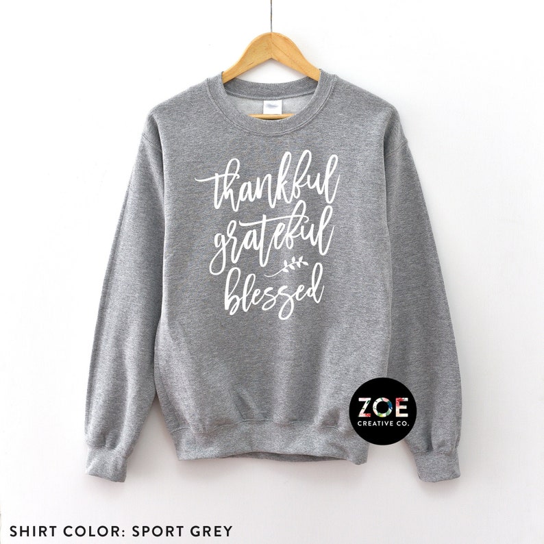 Peut inclure: Un sweat-shirt gris avec le texte "thankful grateful blessed" en lettres blanches. Le sweat-shirt a un col rond et des manches longues. Le texte est &eacute;crit dans une police d'&eacute;criture manuscrite. Le sweat-shirt est sur un fond blanc.