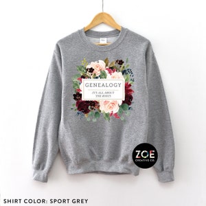 Peut inclure: Un sweat-shirt gris avec un motif floral et le texte "GENEALOGY IT'S ALL ABOUT THE ROOTS" dans un rectangle blanc. Le texte "ZOE CREATIVE CO." est dans le coin inf&eacute;rieur droit.