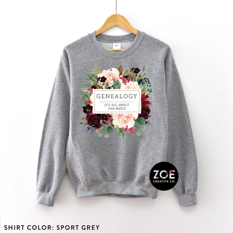 Peut inclure: Un sweat-shirt gris avec un motif floral et le texte "GENEALOGY IT'S ALL ABOUT THE ROOTS" dans un rectangle blanc. Le texte "ZOE CREATIVE CO." est dans le coin inf&eacute;rieur droit.
