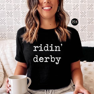May include: Black t-shirt with white text that reads "ridin' derby".