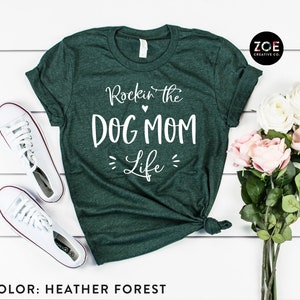 May include: A green heather forest t-shirt with the text "Rockin' the Dog Mom Life" in white. The shirt is laid flat on a white wooden surface with a pair of white sneakers and a bouquet of pink and white roses.