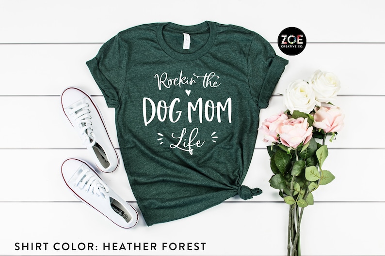 May include: A green heather forest t-shirt with the text "Rockin' the Dog Mom Life" in white. The shirt is laid flat on a white wooden surface with a pair of white sneakers and a bouquet of pink and white roses.