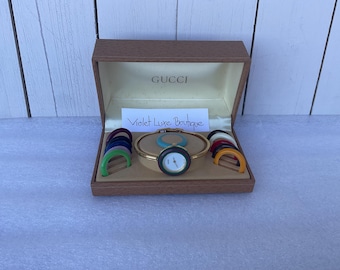 May include: A Gucci watch set in a brown box. The watch has a gold band and a black face with white hands. The set includes interchangeable colored bezels and a turquoise accent. The box has the Gucci logo and a note that says "Violet Luxe Boutique."