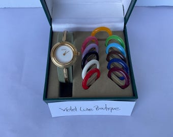 May include: A gold watch with a white face and a green leather band in a box with a variety of colorful plastic bangles. The box has a label that reads "Violet Luxe Boutique".
