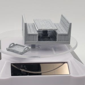 May include: A miniature gray picnic table with attached benches and a small serving tray. The table has a textured wood grain appearance. The set is displayed on a clear, circular platform.