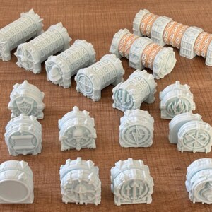 May include: A collection of white 3D-printed cylindrical and semi-cylindrical game components. Several pieces feature rotating number dials with orange or white digits. Other pieces display embossed symbols including shields, flames, and lightning bolts.