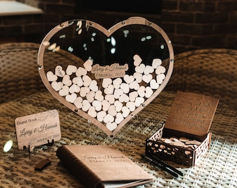 May include: A wooden heart-shaped box with a glass front filled with small wooden hearts. The box is on a table with a guest book, a wooden box with a sign that says "Wedding Guest Book Please sign - A Heart" and two pens.