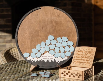 May include: A wooden wedding guest book with a circular, brown surface and a black frame. It features blue cloud-shaped guest sign-in pieces and a mountain design. Includes a small wooden box and a sign with text.