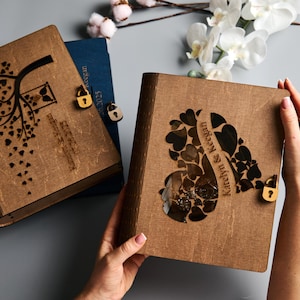 Peut inclure: Deux albums photo en bois avec des motifs de cœur et d'arbre. L'un est ouvert, révélant une découpe en forme de cœur. L'autre présente un motif d'arbre. Les deux ont un texte personnalisé et de petits cadenas en bois.
