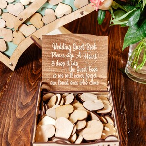 K&ouml;nnte beinhalten: Holzkiste mit einem Schild mit der Aufschrift "Wedding Guest Book Please sign A Heart & drop it into the Guest Book so we will never forget our loved ones who came". Die Kiste ist mit Holzh Herzen gef&uuml;llt.