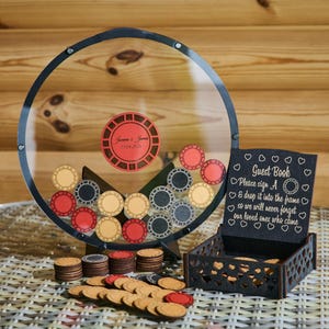 May include: A clear, circular frame with a black border, filled with poker chips in red, black, and tan. A small black box with a sign that reads "Guest Book" is next to the frame. Additional wooden poker chips are scattered around.