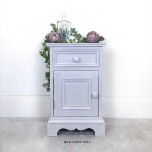 May include: A small, lavender-coloured bedside table with a single drawer and a cabinet door. The table has a decorative base and features a knob on the drawer and door. The table is decorated with a glass bottle and artificial flowers.